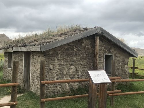 A small, rustic building with a grass-covered roof, surrounded by a wooden fence and a sign, set against a cloudy sky.