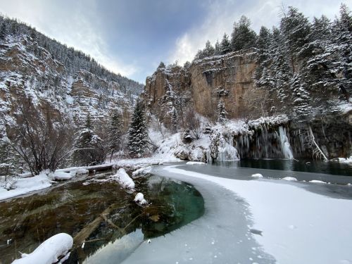 A snowy landscape featuring a river, frozen sections, and towering cliffs surrounded by evergreen trees.