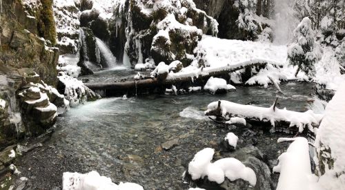 A serene winter scene featuring a snowy landscape with a flowing stream and frozen waterfalls surrounded by rocks.