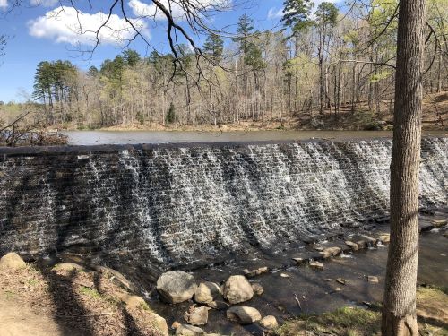 A serene waterfall cascading over a stone dam, surrounded by trees and a calm lake under a clear blue sky.