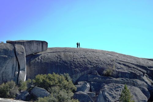 Two people stand on a large rock formation against a clear blue sky, surrounded by trees and boulders.