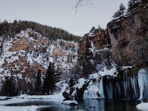 A serene winter landscape featuring a frozen waterfall, snow-covered cliffs, and evergreen trees.