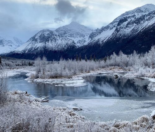 A serene winter landscape featuring a frozen river, snow-covered mountains, and frosted trees under a cloudy sky.