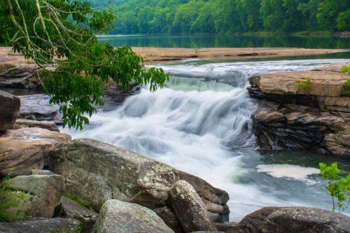 A serene waterfall cascades over rocky ledges, surrounded by lush green trees and a calm river.