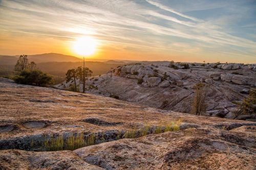 Sunset over rocky terrain, with sparse vegetation and distant hills under a colorful sky.