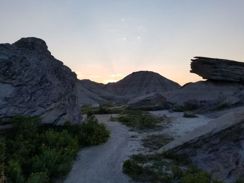 A rocky landscape at sunset, with hills in the background and a path leading through the foreground vegetation.