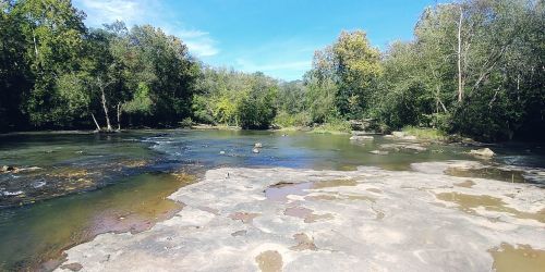 A serene river scene with rocky banks, surrounded by lush green trees under a clear blue sky.