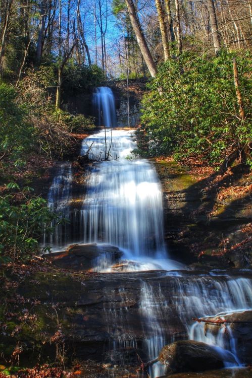 A serene waterfall cascades down rocky steps, surrounded by lush greenery and trees under a clear blue sky.