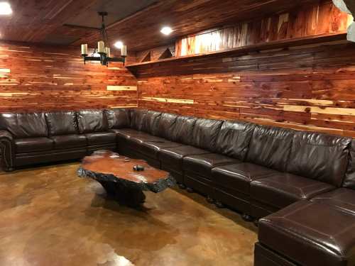 Cozy living room with a large brown leather sectional sofa and a rustic wooden coffee table against a wooden wall.