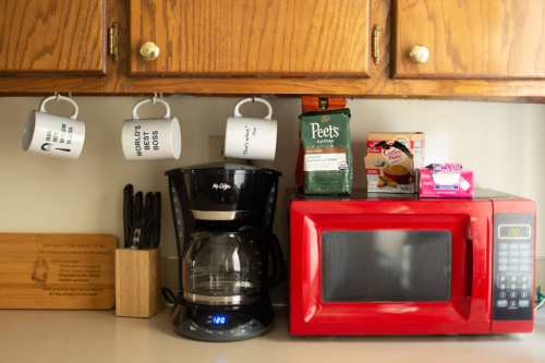 A kitchen counter with a coffee maker, microwave, coffee boxes, and hanging mugs on wooden cabinets above.