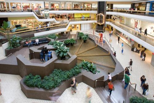 A spacious mall interior with multiple levels, greenery, a water feature, and shoppers walking around.
