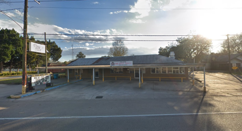 A small, single-story building with a metal roof and a sign, set against a cloudy sky and empty parking lot.