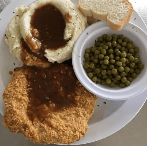 A plate with fried chicken smothered in gravy, mashed potatoes, green peas, and a slice of bread.