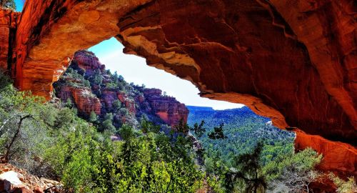 A stunning view of a red rock arch framing a lush green valley and distant hills under a clear blue sky.