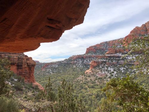 A scenic view of red rock formations and lush greenery under a cloudy sky, framed by a rocky overhang.