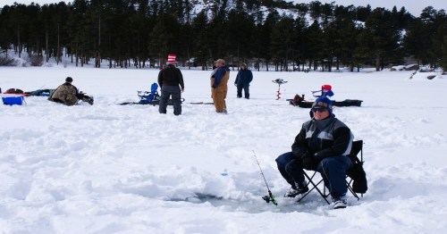 A group of people ice fishing on a snowy lake, with one person sitting on a chair by a hole in the ice.