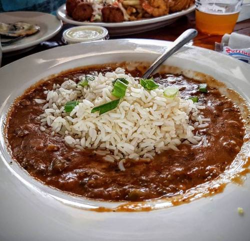 A bowl of rich brown stew topped with white rice and green onions, served on a plate with other dishes in the background.