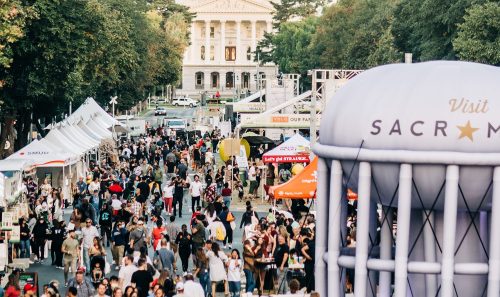 A bustling outdoor event in Sacramento, with crowds, food stalls, and a historic building in the background.