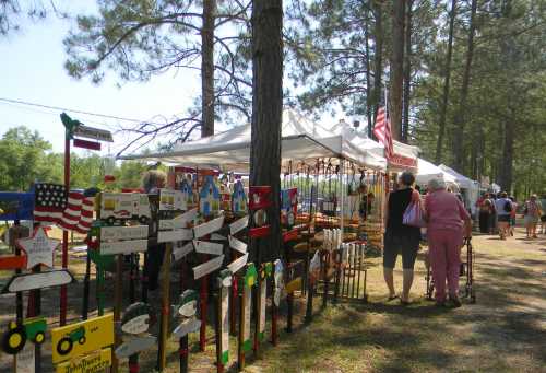 A bustling outdoor market with vendors under tents, surrounded by trees and visitors browsing colorful signs and crafts.