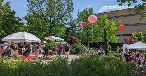 A lively outdoor scene with people at tables, colorful balloons, and greenery near a building on a sunny day.