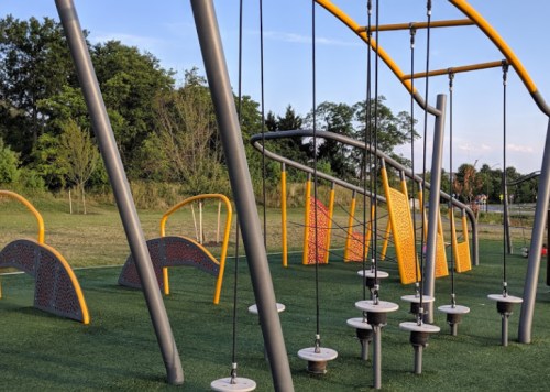 A modern playground with swings and climbing structures on green turf, surrounded by trees and open space.