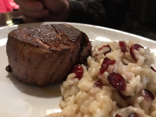 A perfectly cooked steak beside a serving of creamy risotto with cranberries on a white plate.