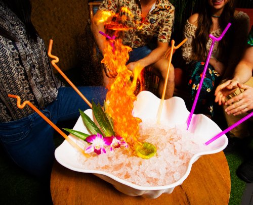 A vibrant drink with flames rising from it, surrounded by ice and colorful straws, with friends enjoying the moment.