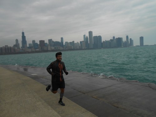 A person jogs along a waterfront path with a city skyline in the background on a cloudy day.