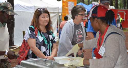 A food vendor serves a meal to customers at an outdoor event, with tents and people in the background.