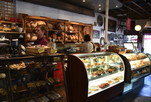 A cozy bakery interior with shelves of bread, a display case of pastries, and two staff members serving customers.