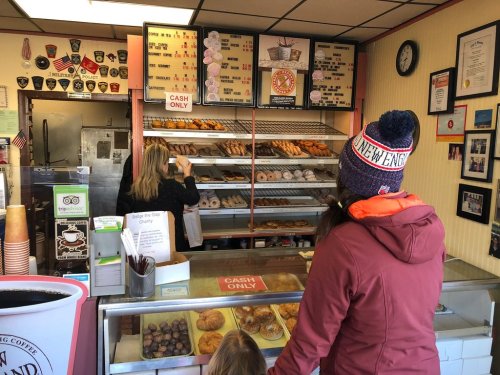 A woman in a winter hat stands at a bakery counter, looking at a display of donuts and pastries. Cash only sign visible.