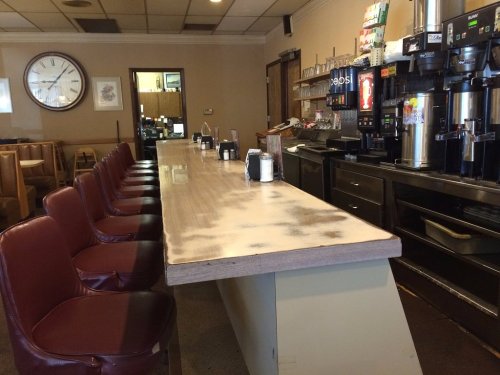 A diner counter with empty maroon chairs, a clock on the wall, and drink dispensers in the background.