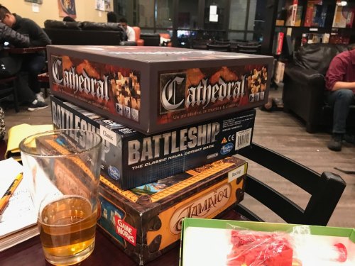 A stack of board games, including "Cathedral," "Battleship," and "Zombicide," on a table with a glass of beer.