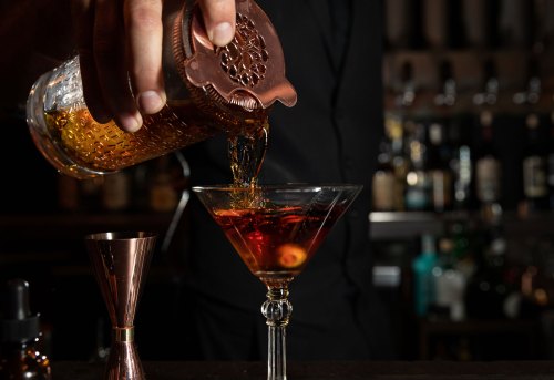 A bartender pours a cocktail from a decorative shaker into a martini glass, with various bottles in the background.