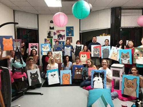 A group of people holding up cat paintings in a colorful room with lanterns and art on the walls.