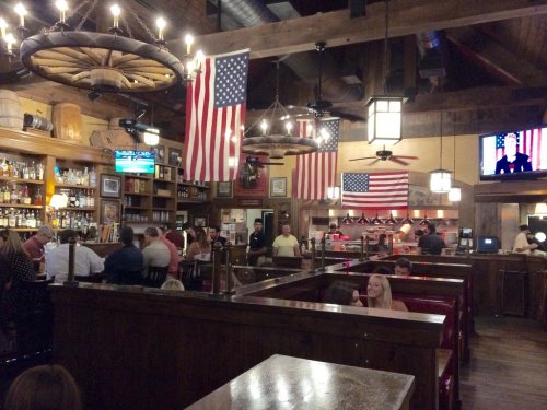 A lively restaurant interior with wooden decor, American flags, and patrons dining at booths and tables.