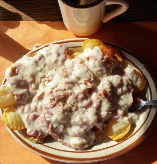 A plate of loaded nachos topped with creamy sauce, diced meat, and black pepper, alongside a cup of coffee.