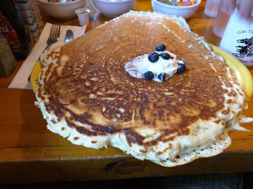 A giant pancake topped with whipped cream and blueberries, served on a wooden table.