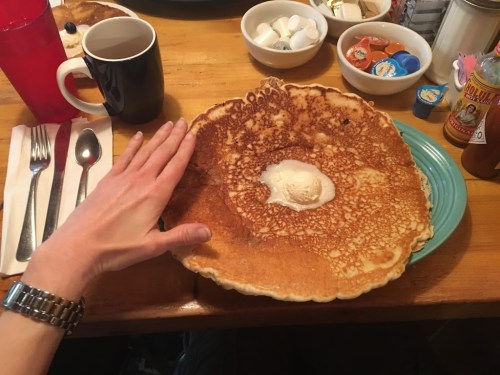 A large pancake with a scoop of butter in the center, with a hand resting on the edge, surrounded by condiments.