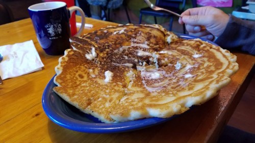 A large, fluffy pancake with melted butter on a blue plate, next to a coffee mug on a wooden table.