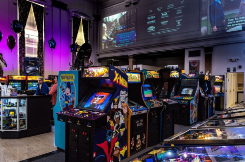 A vibrant arcade filled with classic game machines, colorful decor, and a large screen displaying content in the background.