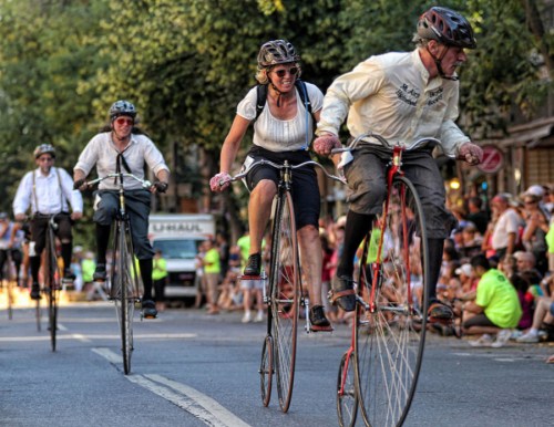 A group of cyclists on vintage high-wheel bicycles ride down a street, with spectators watching from the sidelines.