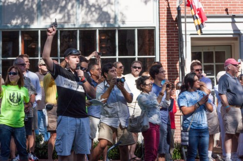 A diverse crowd gathers outdoors, some holding cameras and phones, while one man raises his arm, speaking passionately.