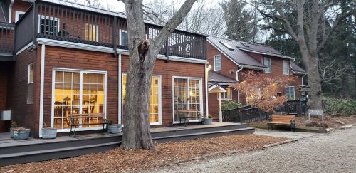 A cozy wooden building with large windows, surrounded by trees and gravel paths, creating a warm, inviting atmosphere.