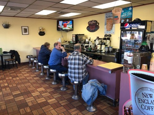 Three customers sit at a counter in a cozy café, with a TV and coffee machines in the background.