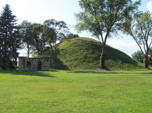 A grassy landscape featuring a large burial mound and a small stone structure under a clear blue sky.