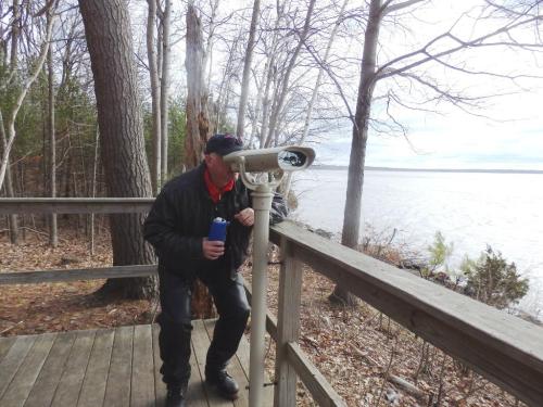 A person leans on a railing, looking through a telescope at a lake surrounded by trees.