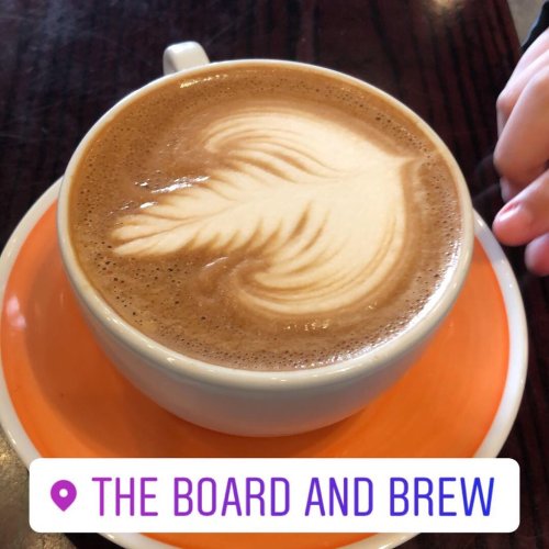 A cup of latte art with a leaf design, placed on an orange saucer at The Board and Brew.