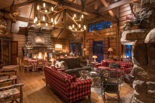 Cozy rustic living room with wooden beams, plaid sofas, a stone fireplace, and warm lighting.