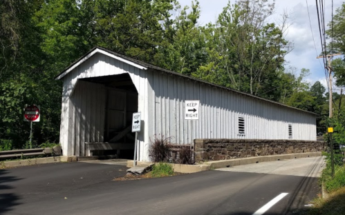 A white covered bridge with a stone foundation, surrounded by trees and road signs, on a sunny day.